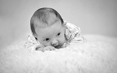 Ein niedliches Kleinkind in Bauchlage auf einem flauschigen, hellen Untergrund, schaut neugierig zur Seite. Das Schwarz-Weiß-Foto betont die Zartheit und Unschuld des Babys. Eine liebevolle Erinnerung, professionell festgehalten von Fotograf Michael Schalansky.